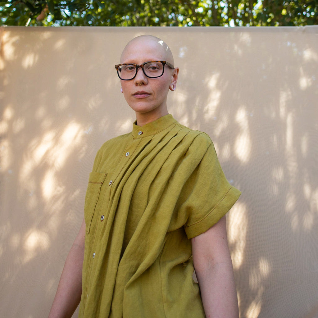 Model standing against a cream color backdrop. Half spin with a half smile wearing Linen, Asymmetrical Pleated Shirt - Chartreuse. Made in California, USA by Rebecca Rae Design Inc. Gender-Inclusive Clothing.
